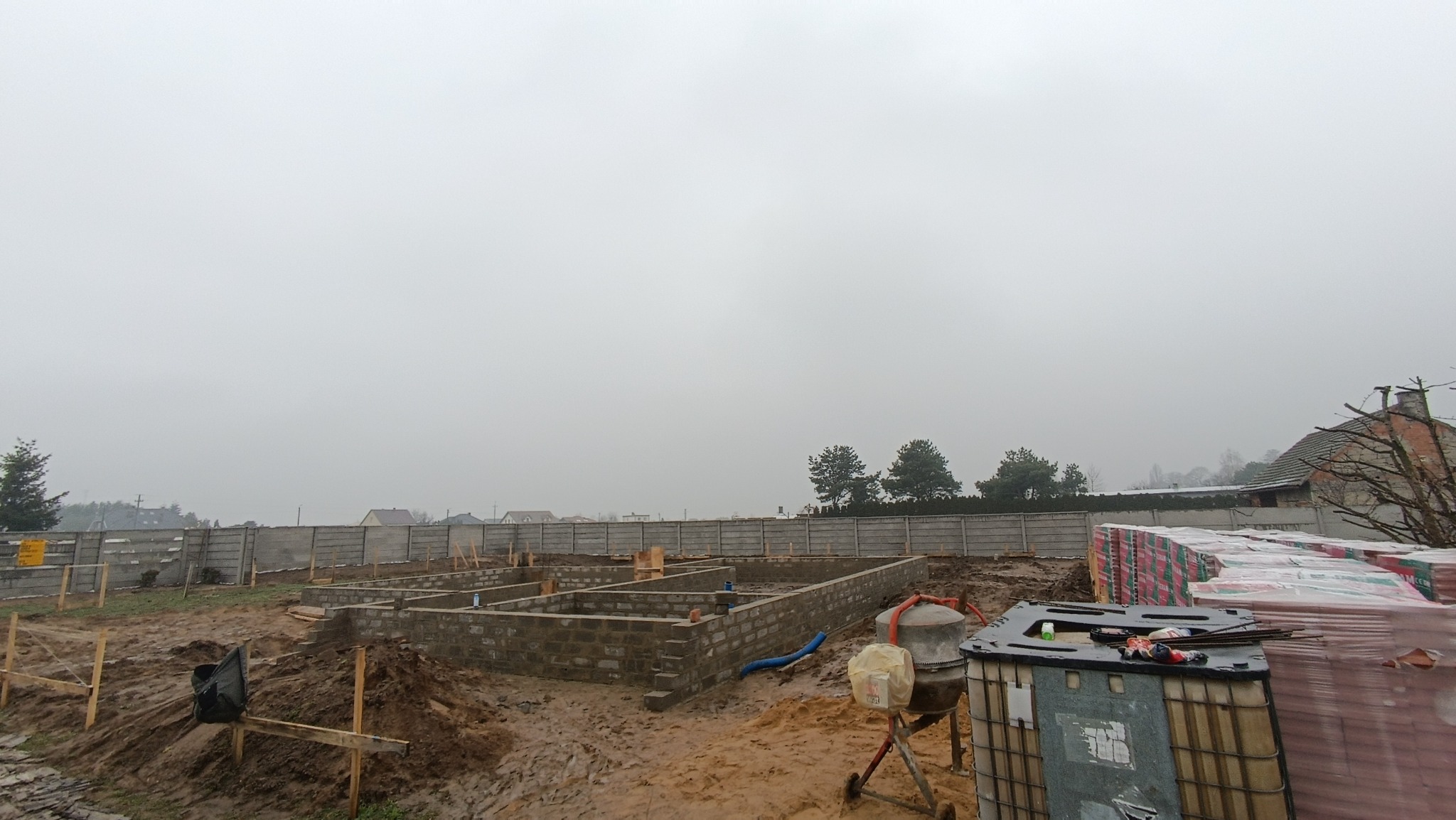 Fundamenty domu z bloczków betonowych na przygotowanym terenie, widoczna betoniarka i materiały budowlane, ogrodzenie w tle, pochmurny dzień.