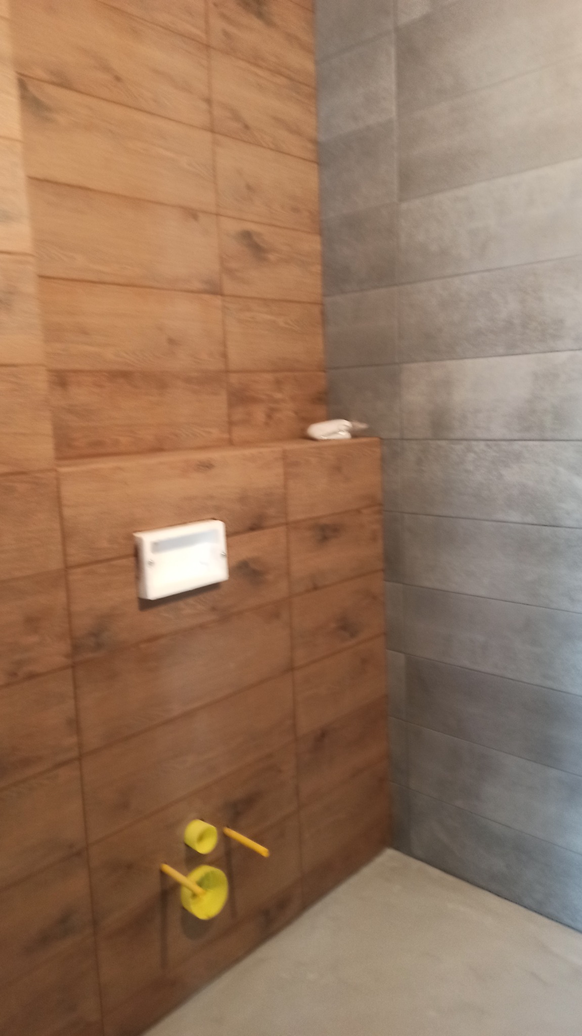 Wykończona łazienka z drewnopodobnymi płytkami na ścianie, szarą płytką na sąsiedniej ścianie, żółte rurki wystające ze ściany oraz biały przycisk spłuczki.