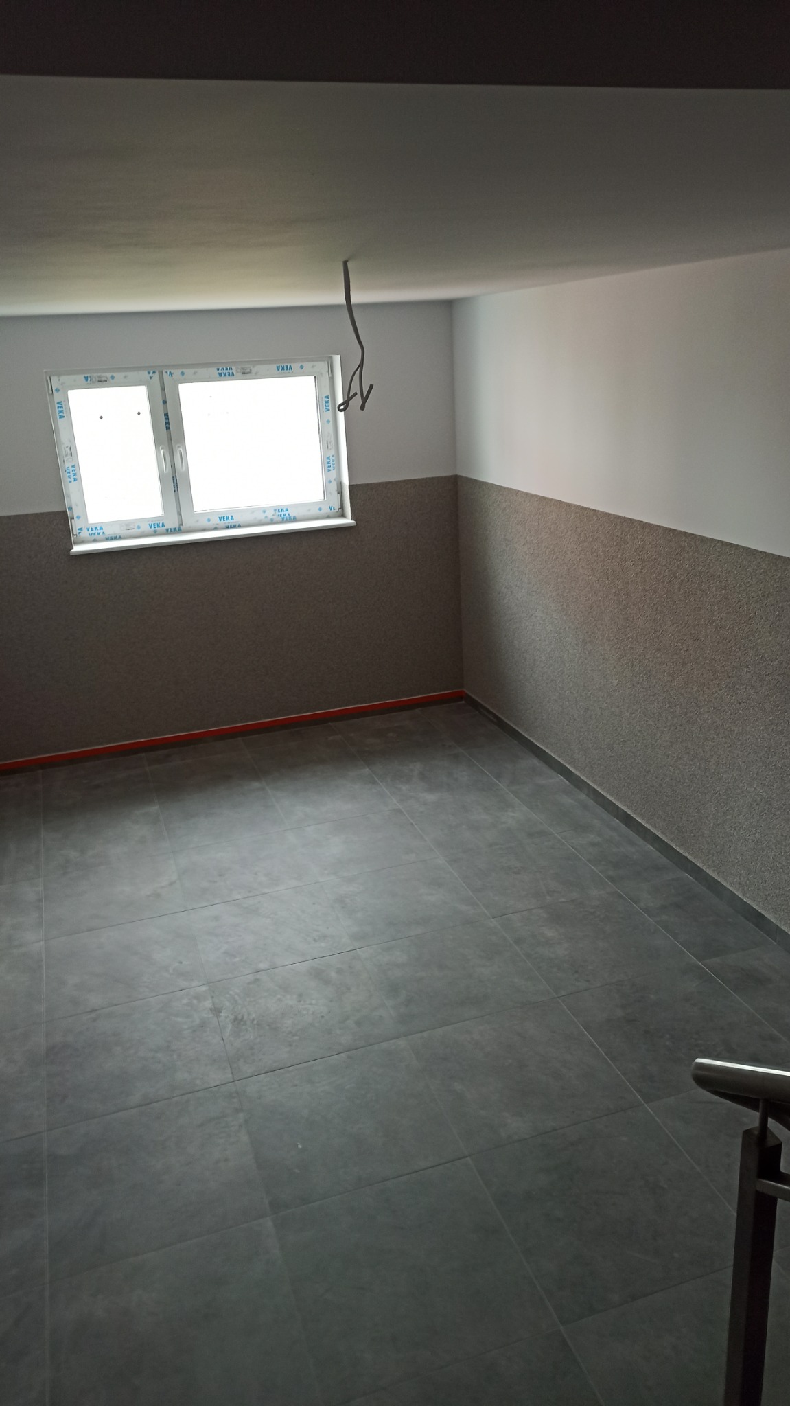Pusty pokój po remoncie z szarymi płytkami podłogowymi, ozdobną tapetą do połowy ściany i wystającymi kablami z sufitu.