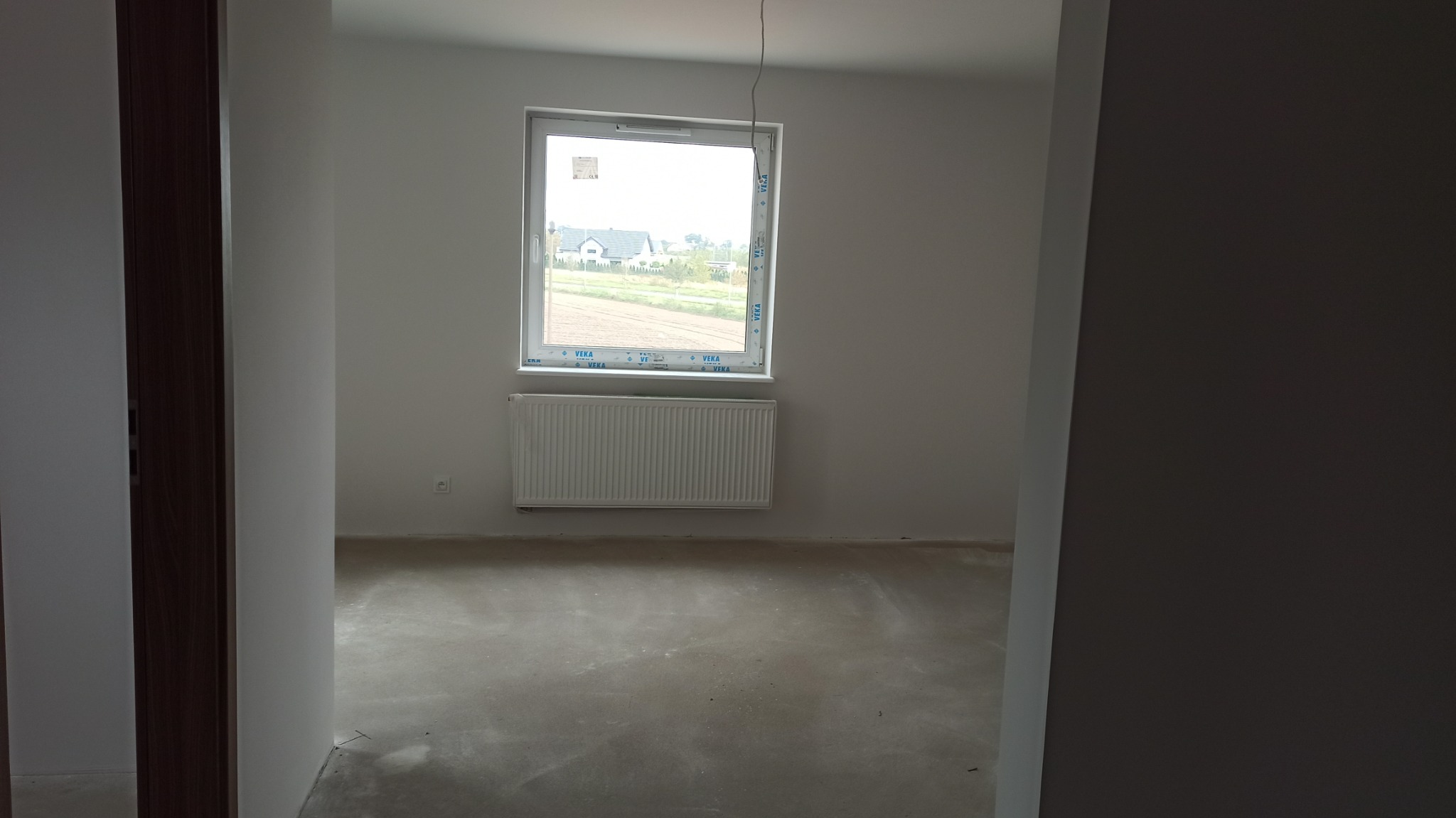Surowe wnętrze nowego domu z oknem na krajobraz i grzejnikiem pod nim, widok przez otwarte drzwi z ciemną futryną.