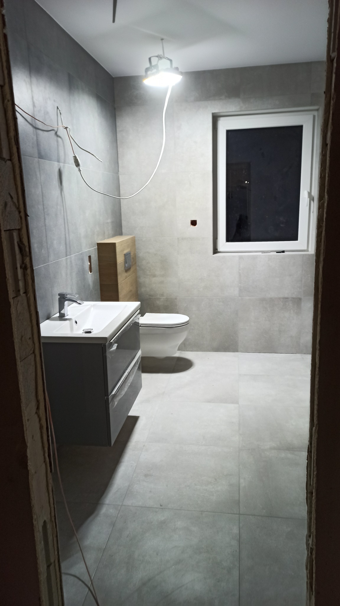 Szara łazienka w trakcie remontu, widoczne płytki, umywalka z szafką, toaleta, okno, tymczasowe oświetlenie i wystające kable.