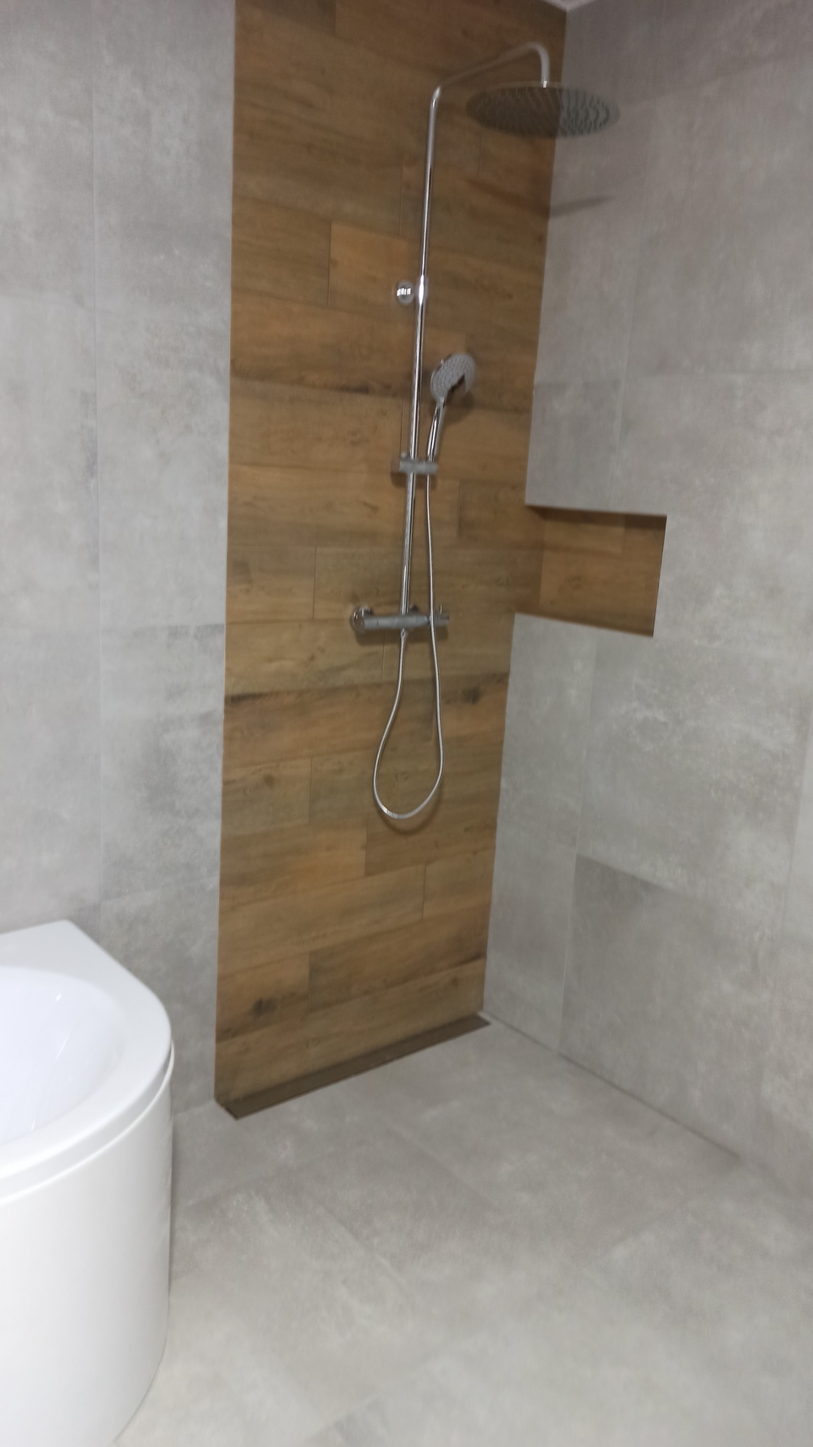 Nowoczesna kabina prysznicowa z deszczownicą, obłożona płytkami imitującymi drewno i szarym kamieniem, z widocznym fragmentem białej toalety.