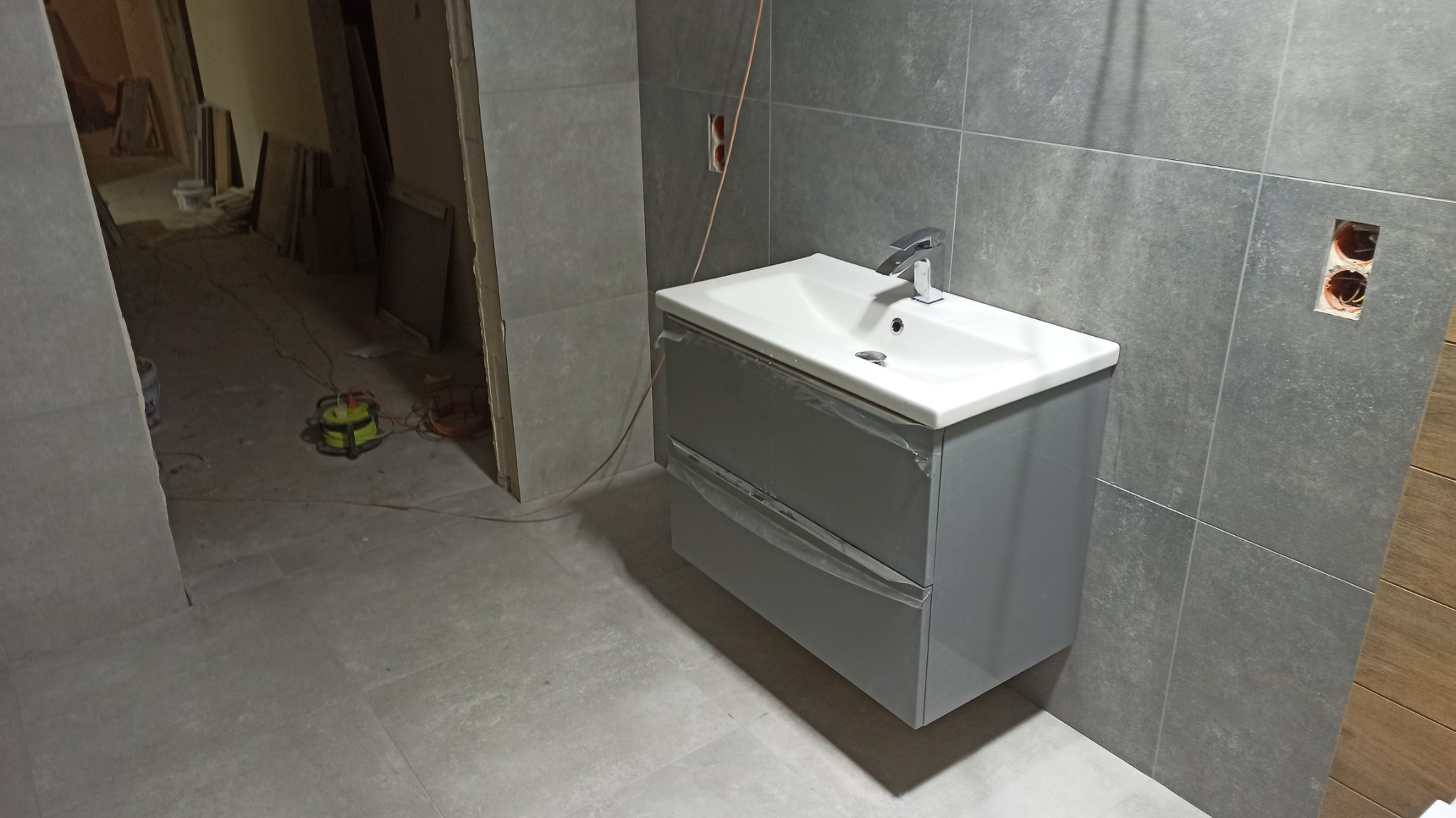 Szara szafka pod umywalkę z białą umywalką i chromowaną baterią, zabezpieczona folią ochronną, w trakcie remontu łazienki z szarymi płytkami, widoczne otwarte gniazdko elektryczne i narzędzia w tle.