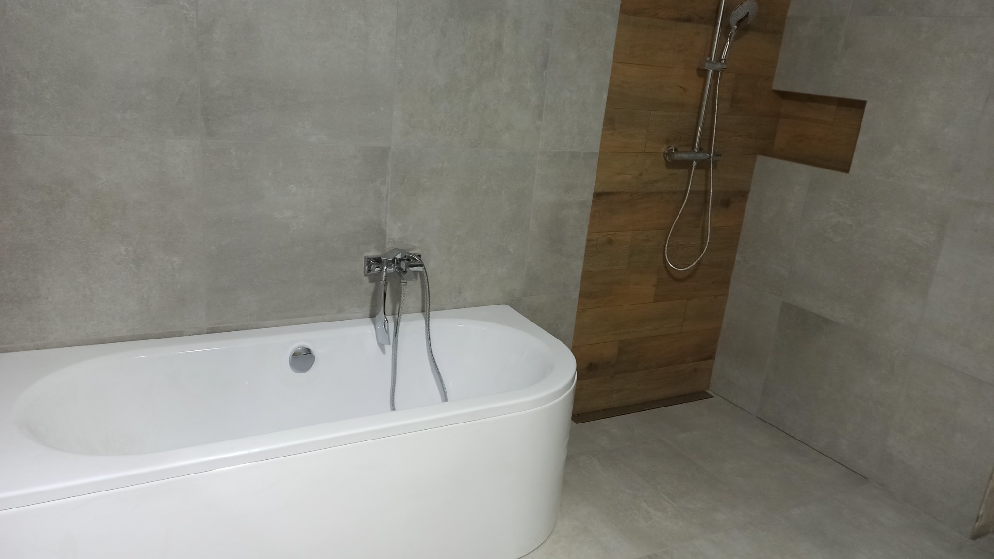 Nowoczesna łazienka z białą wanną, szarą płytką ścienną i drewnianą wstawką przy prysznicu.