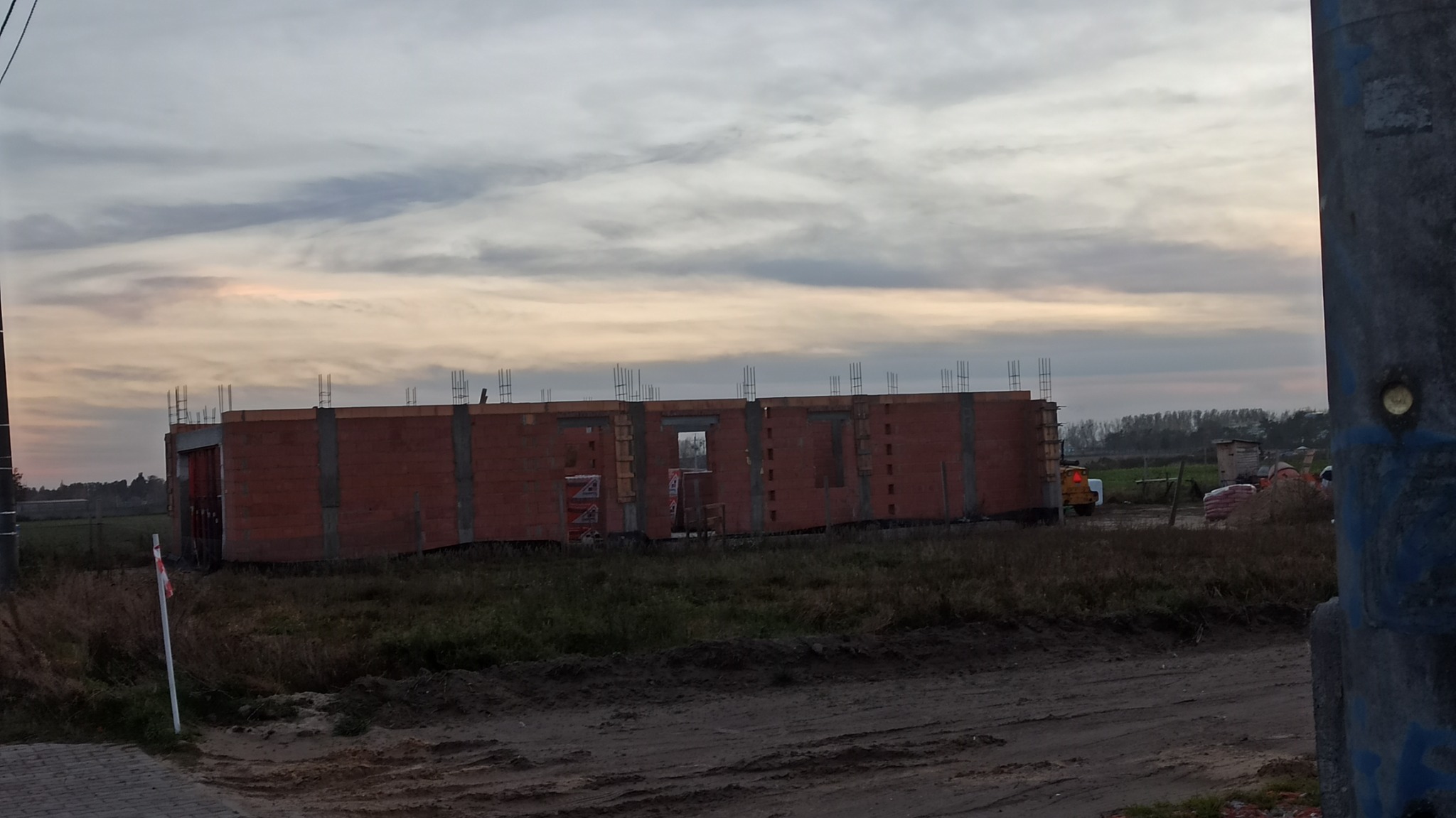 Wznoszony dom murowany z czerwonej cegły, widoczne zbrojenia, teren budowy z piaskiem i gruzem, pochmurne niebo o zmierzchu.