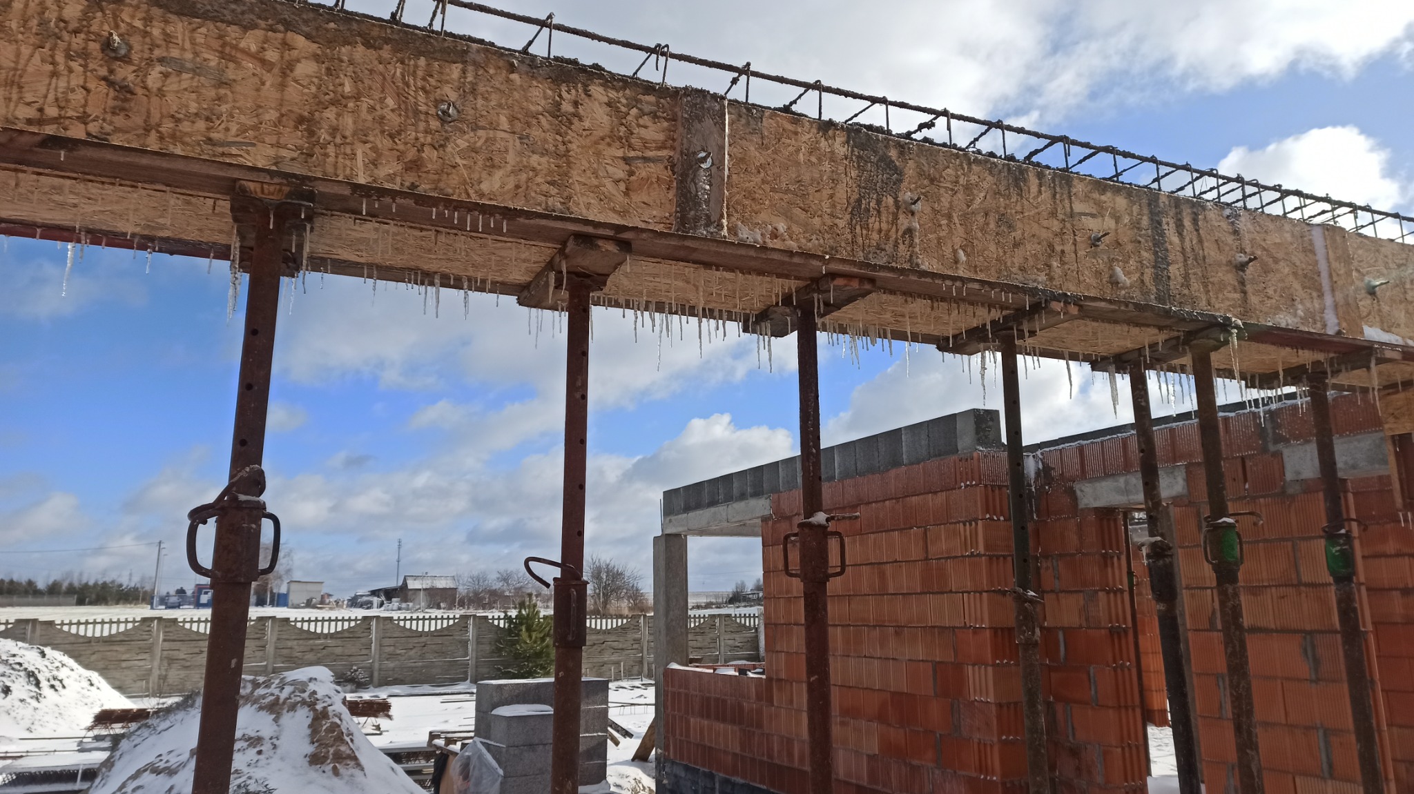 Wznoszenie murów domu z czerwonej cegły z widocznym szalunkiem i podporami stalowymi, z wiszącymi soplami lodu w słoneczny dzień.