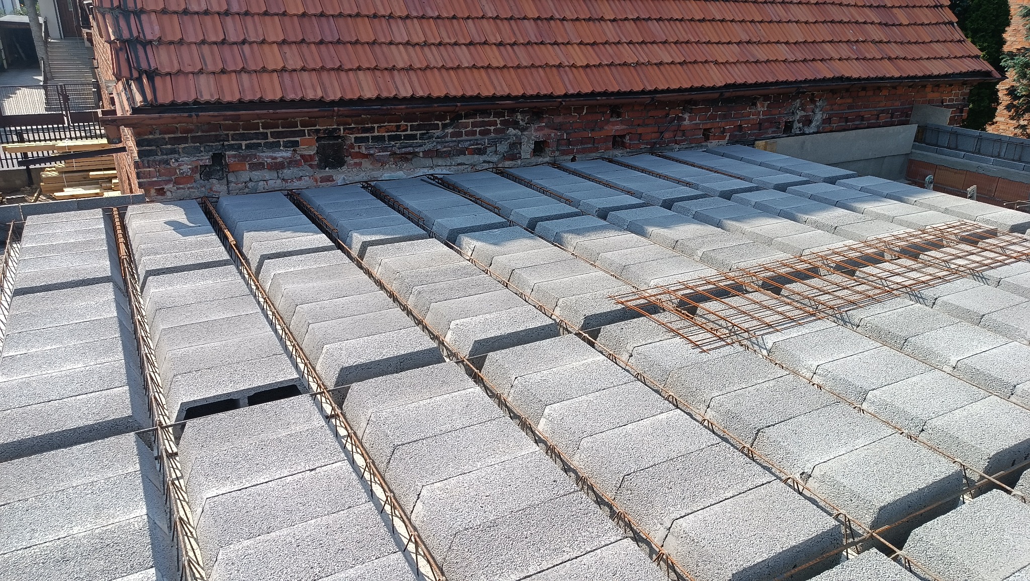 Wieniec stropowy Teriva w trakcie montażu, z widocznym zbrojeniem i bloczkami szalunkowymi, na tle czerwonej cegły i dachu z dachówki ceramicznej.