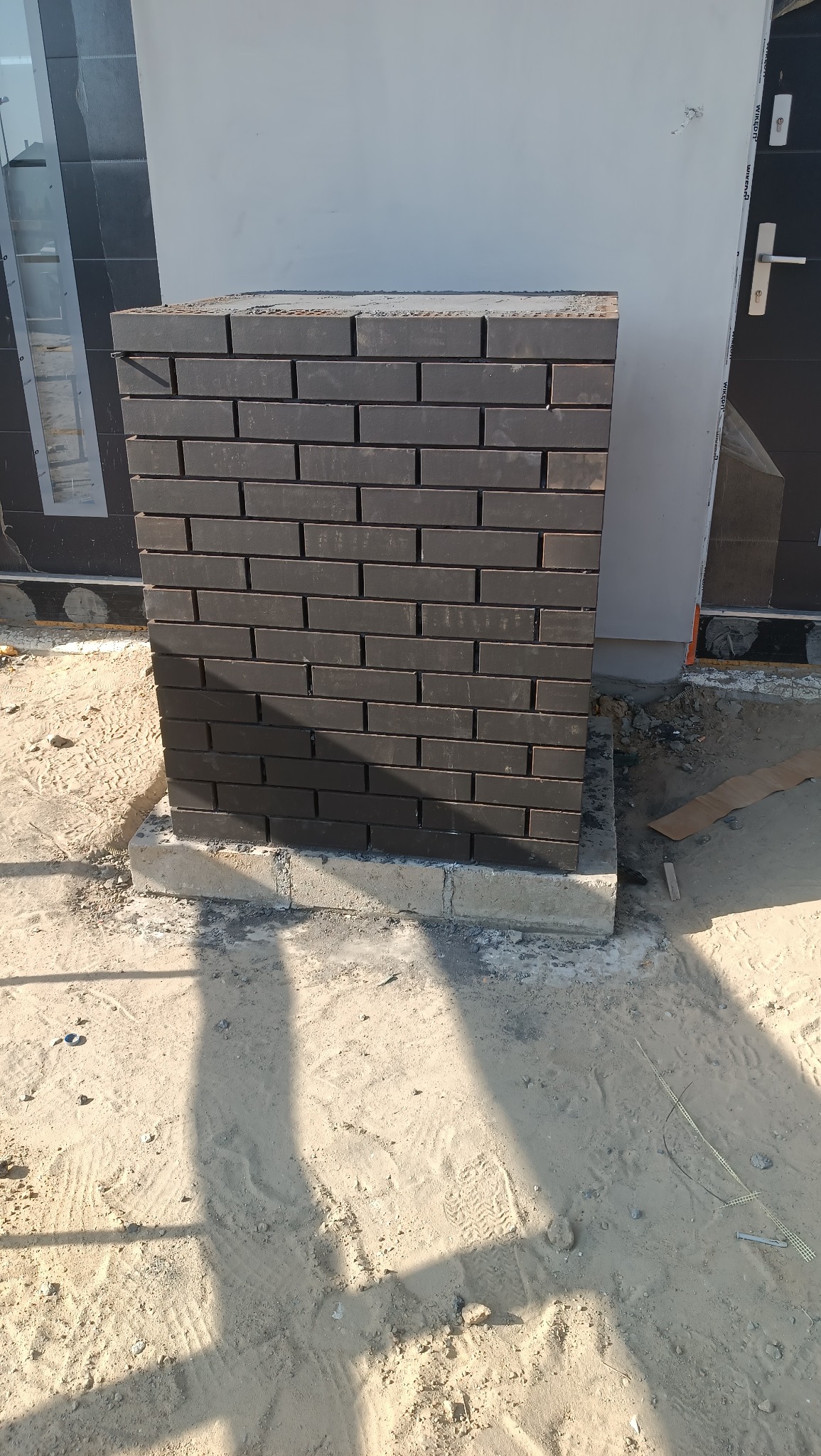 Nowo wymurowany, ciemnobrązowy filar z cegły klinkierowej na betonowej podstawie, przy elewacji budynku w trakcie budowy, z widocznym piaskiem i elementami budowlanymi.