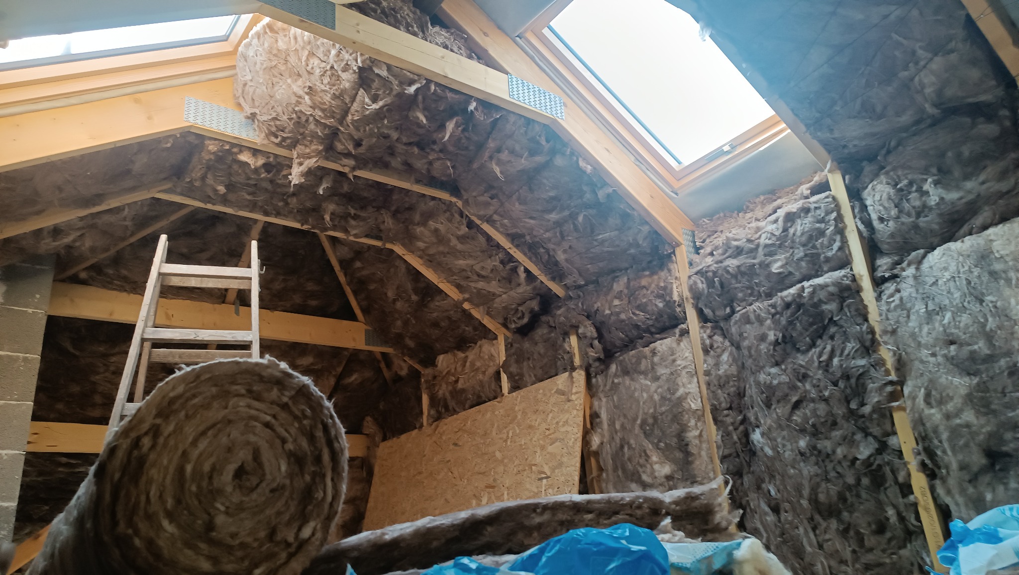 Poddasze w trakcie ocieplania wełną mineralną, widoczna drewniana konstrukcja dachu, okno dachowe i drabina oparta o ścianę.