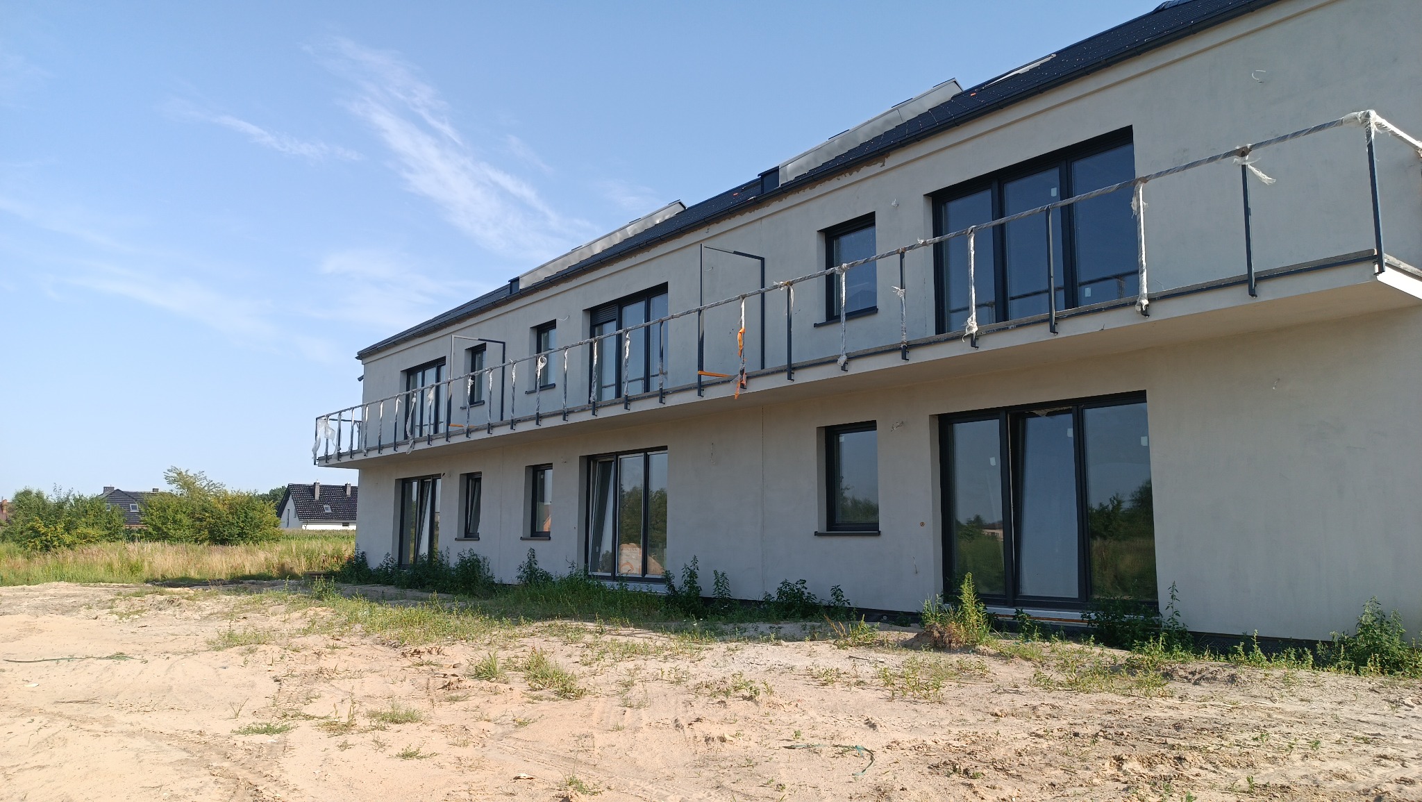 Dwukondygnacyjny dom murowany w trakcie budowy z czarną dachówką i balkonami z metalowymi balustradami zabezpieczonymi folią, widok z poziomu gruntu na niezagospodarowaną działkę.