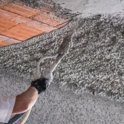 Aplikacja szarego tynku maszynowego na ścianie z widocznymi cegłami na górze, wykonywana przez pracownika w rękawicy i białej koszulce przy użyciu specjalistycznego pistoletu natryskowego.