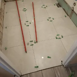 Układanie jasnych, marmurkowych płytek podłogowych z użyciem poziomnic i klipsów poziomujących w łazience w Skalbmierzu, widoczny fragment podłogi z panelami imitującymi drewno.