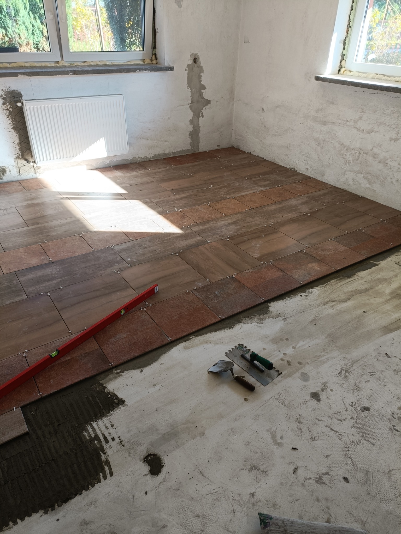 Proces układania brązowych płytek podłogowych z użyciem poziomicy, szpachelek i krzyżyków dystansowych w pomieszczeniu z surowymi ścianami i grzejnikiem pod oknem.