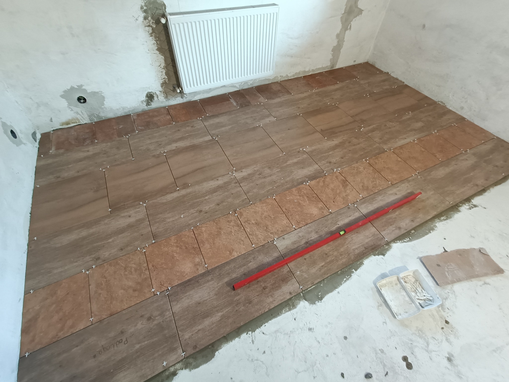 W trakcie układania podłogi z płytek imitujących drewno, widoczne krzyżyki dystansowe i poziomica, surowe ściany w tle.