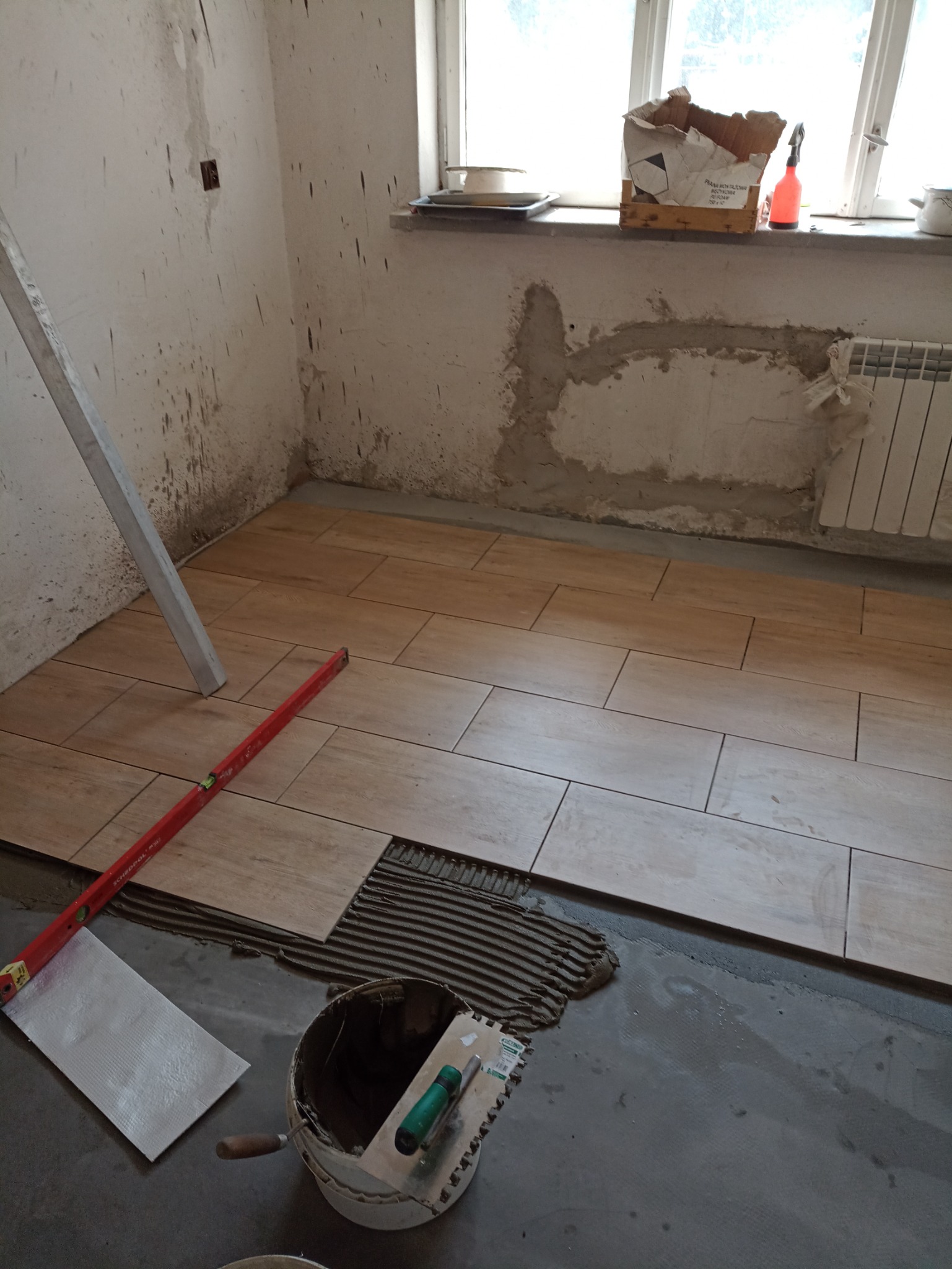 Układanie podłogi z płytek imitujących drewno, widoczny poziomica, kielnia, wiadro z klejem i fragment ściany w stanie surowym.