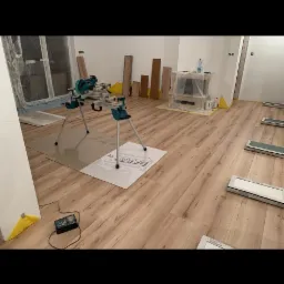 Układanie paneli podłogowych w jasnym wnętrzu, widoczna piła ukośnica na stojaku, panele ułożone częściowo, zabezpieczone folią meble w tle.