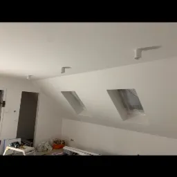 Poddasze ze świeżo pomalowanymi na biało ścianami i sufitem, widoczne okna dachowe zabezpieczone folią malarską, surowe, minimalistyczne oświetlenie punktowe, częściowo zagracone narzędziami...