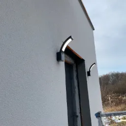 Dwa nowoczesne, czarne kinkiety w kształcie łuku zamontowane na świeżo otynkowanej, białej elewacji budynku w trakcie wykończenia, widoczny fragment nieobrobionego otworu drzwiowego i drabina w tle.