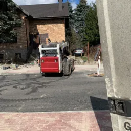 Prace brukarskie w toku: miniładowarka na asfalcie, w tle budynek z cegły i pracownik przygotowujący podłoże pod kostkę, widoczny fragment nowej nawierzchni z czerwonej kostki.