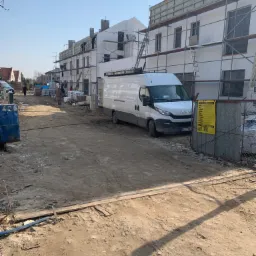 Budowa szeregu domów w trakcie prac elewacyjnych, widoczne rusztowania, materiały budowlane i biały samochód dostawczy na placu budowy.