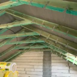 Konstrukcja dachu z impregnowanego drewna z metalowymi mocowaniami, widoczna izolacja termiczna w rolkach i mur z bloczków, pracownik siedzący na dole sprawdza postęp prac.