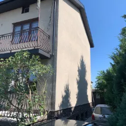 Fragment elewacji budynku z balkonem i widocznym fragmentem ocieplenia przy gruncie, z metalową anteną na dachu i cieniem drzewa na ścianie.