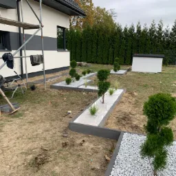 Nowo założony ogród z roślinami w geometrycznych rabatach wyłożonych białym grysem, na tle domu z szarymi akcentami i zielonych tui.