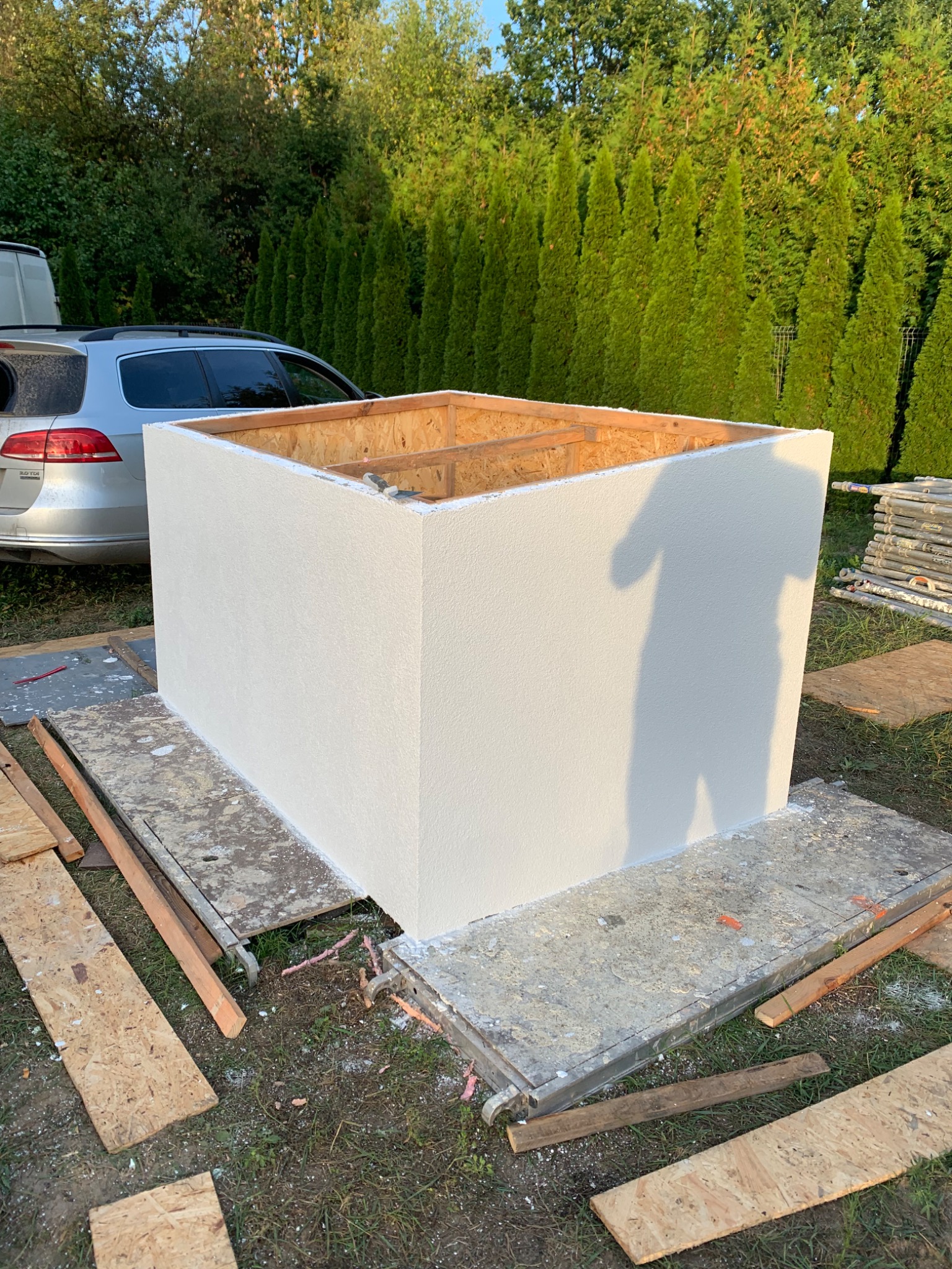 Biała, otynkowana konstrukcja w kształcie sześcianu, z widoczną drewnianą ramą od góry, ustawiona na platformie roboczej na trawie, z samochodem i żywopłotem w tle.