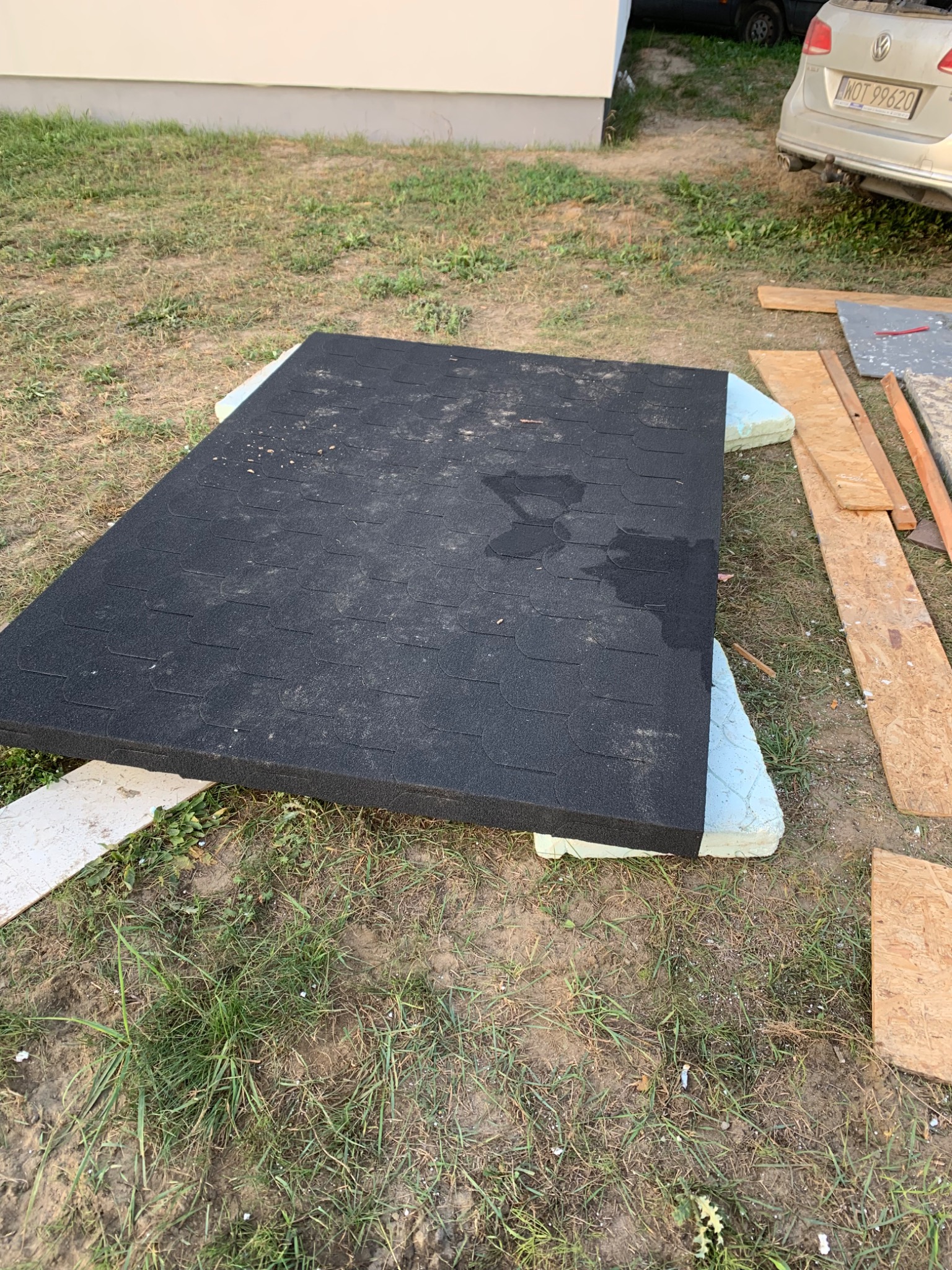 Czarny arkusz papy termozgrzewalnej na styropianowych podkładkach, składowany na trawie obok desek i zaparkowanego samochodu.