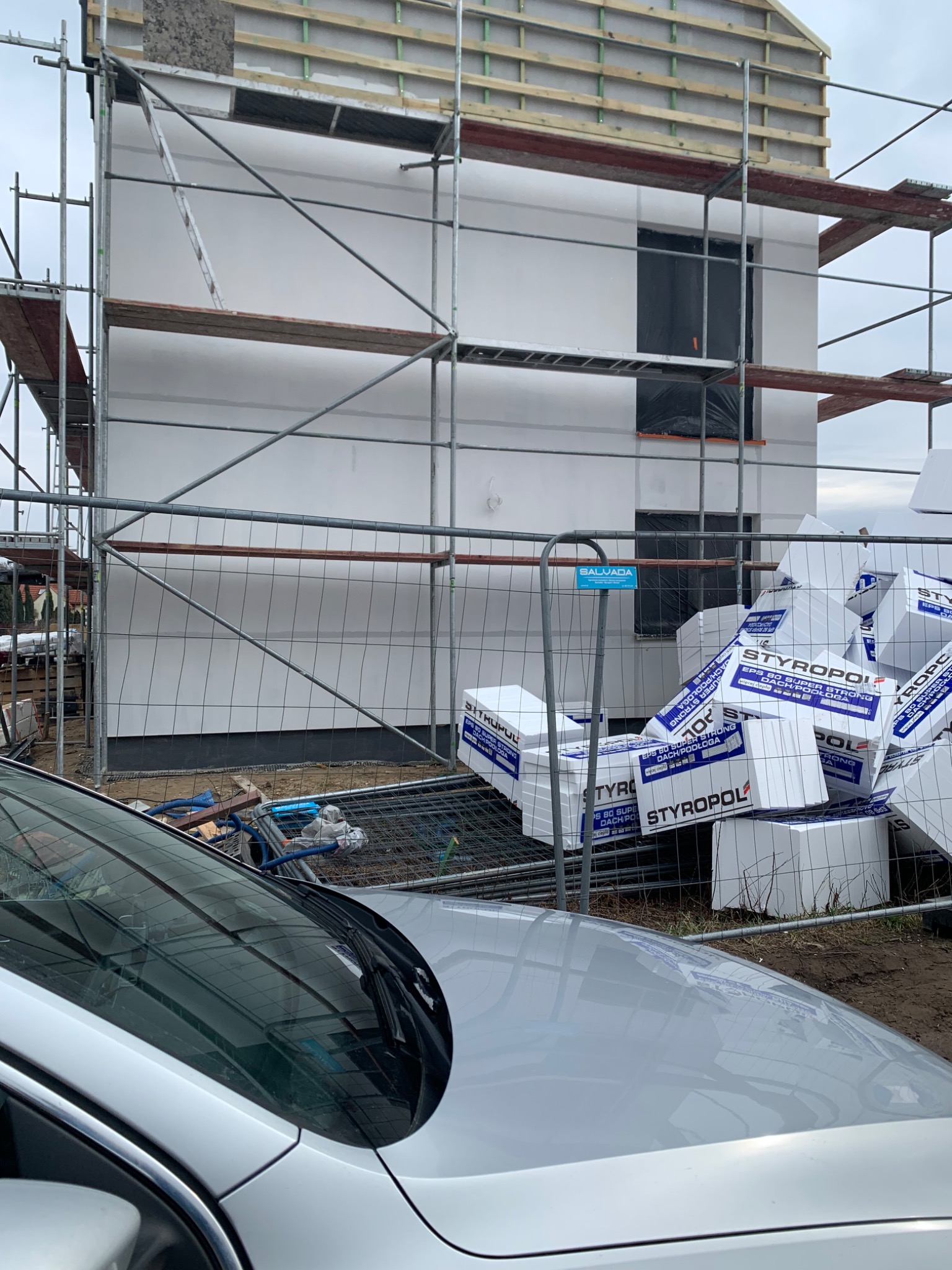 Dom w trakcie ocieplania, widoczne rusztowania i stos paczek styropianu. W tle fragment srebrnego samochodu.