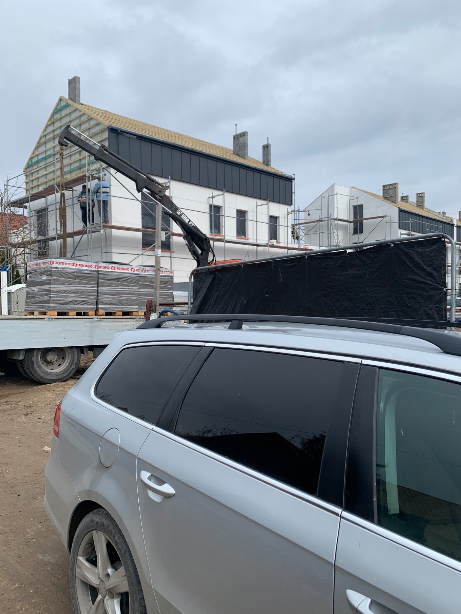 Montaż elewacji na nowym budynku z użyciem dźwigu, widoczne rusztowania i palety z materiałem budowlanym. W tle zaparkowany samochód osobowy.