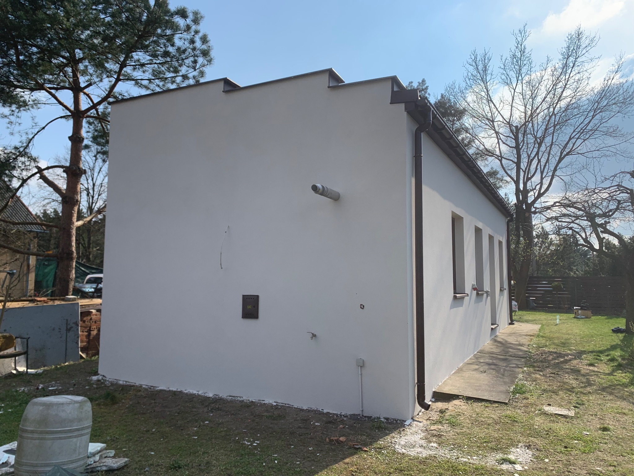 Świeżo wykończona, jasnoszara elewacja budynku z brązowymi rynnami, widoczny fragment instalacji gazowej na ścianie, drzewa w tle.
