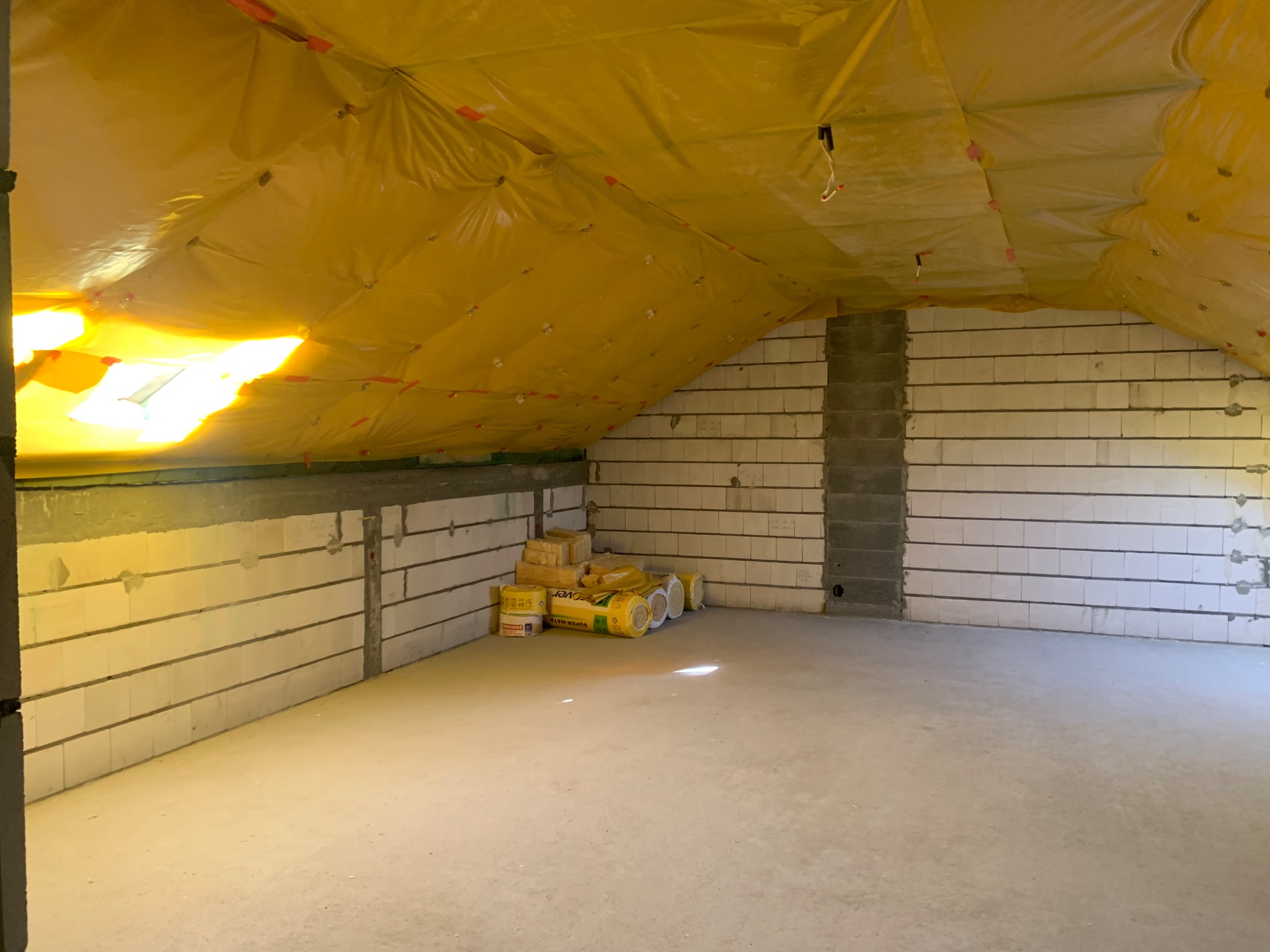 Poddasze z ociepleniem z folii i wełny mineralnej, bloczki betonowe na ścianach, częściowo otynkowane, widoczne rolki wełny mineralnej w opakowaniach.