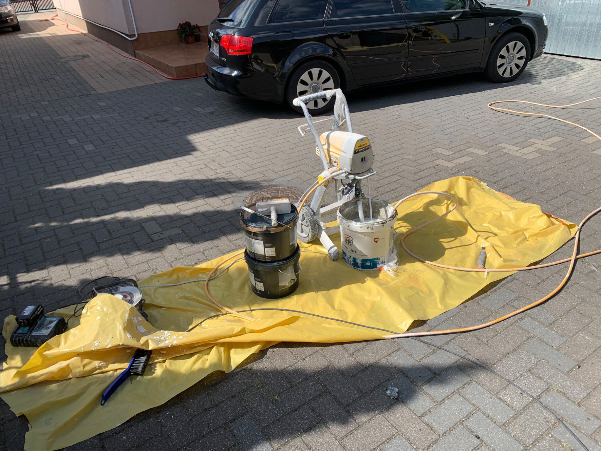 Sprzęt do malowania natryskowego, dwie puszki farby i akcesoria rozłożone na żółtej folii ochronnej na kostce brukowej, w tle czarny samochód.