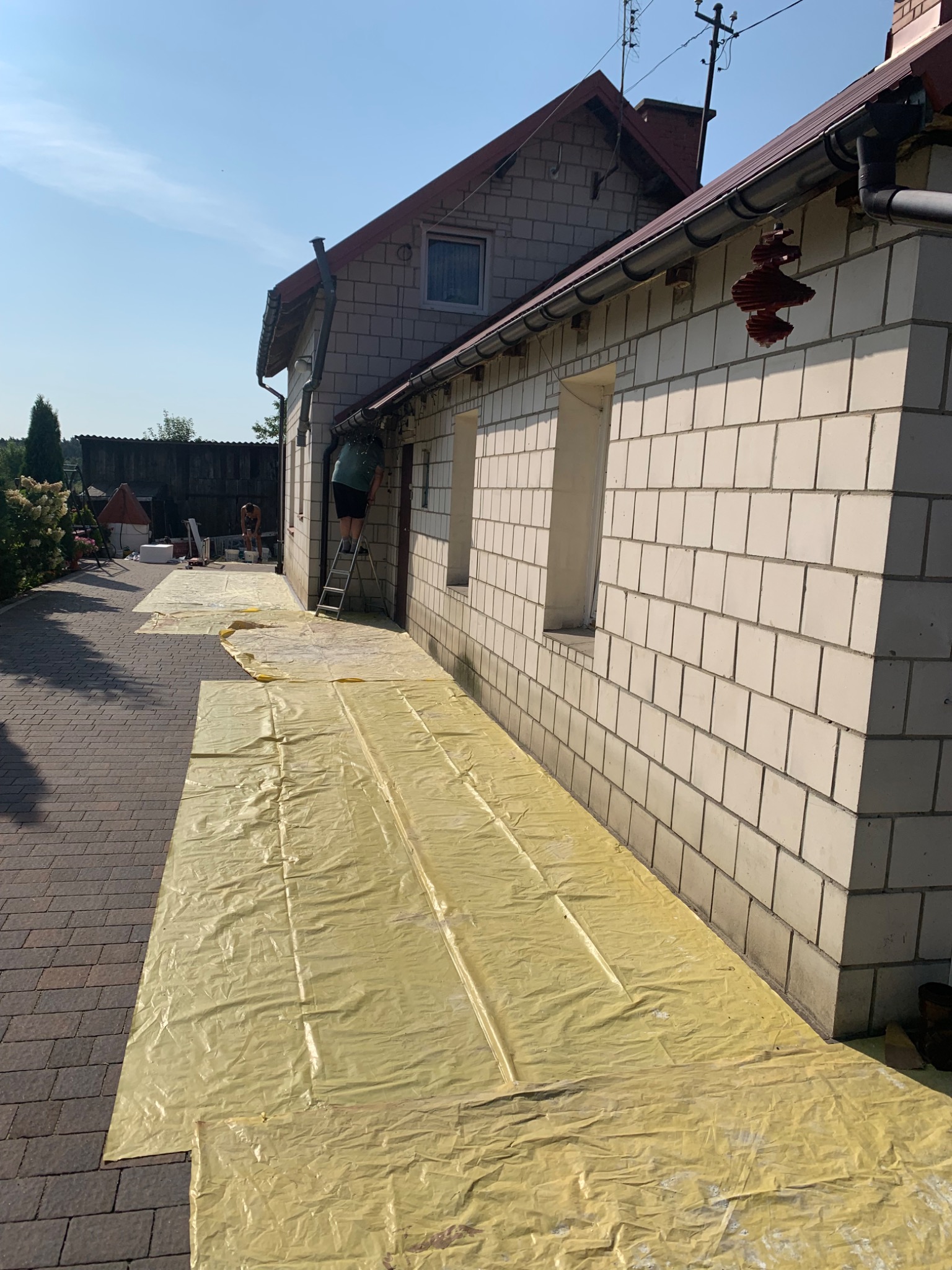 Przygotowanie do prac elewacyjnych: rozłożona folia ochronna na brukowanym podłożu, pracownik na drabinie przy ścianie budynku z białej cegły, drugi pracownik w tle.