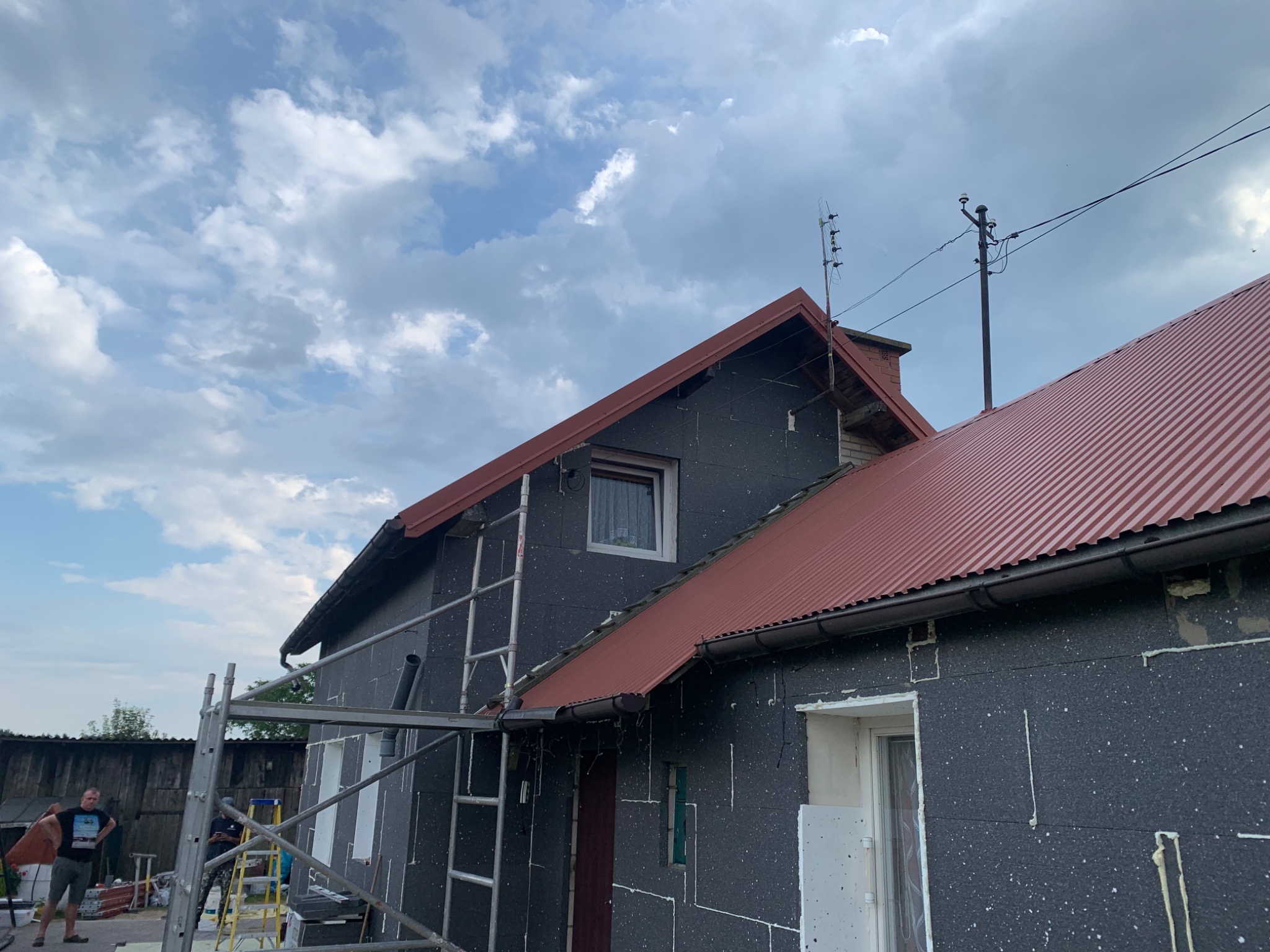 Dom w trakcie ocieplania, widoczne szare płyty styropianowe na elewacji, czerwony dach z blachy trapezowej, rusztowanie i drabina oparta o ścianę, pochmurne niebo w tle.