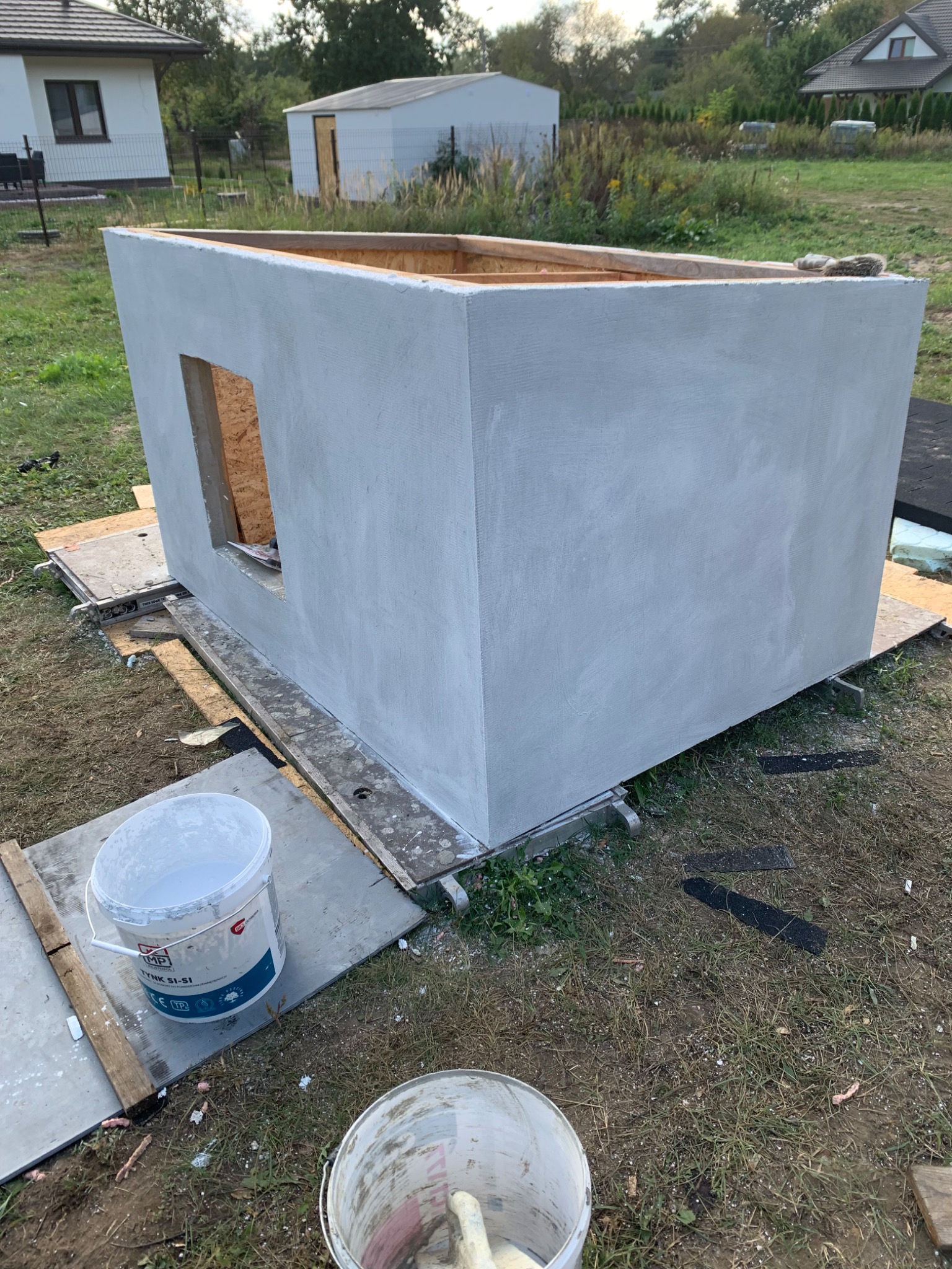 Szara konstrukcja budynku z otworem okiennym, w trakcie prac wykończeniowych, widoczne wiaderka z farbą i elementy rusztowania na zewnątrz.