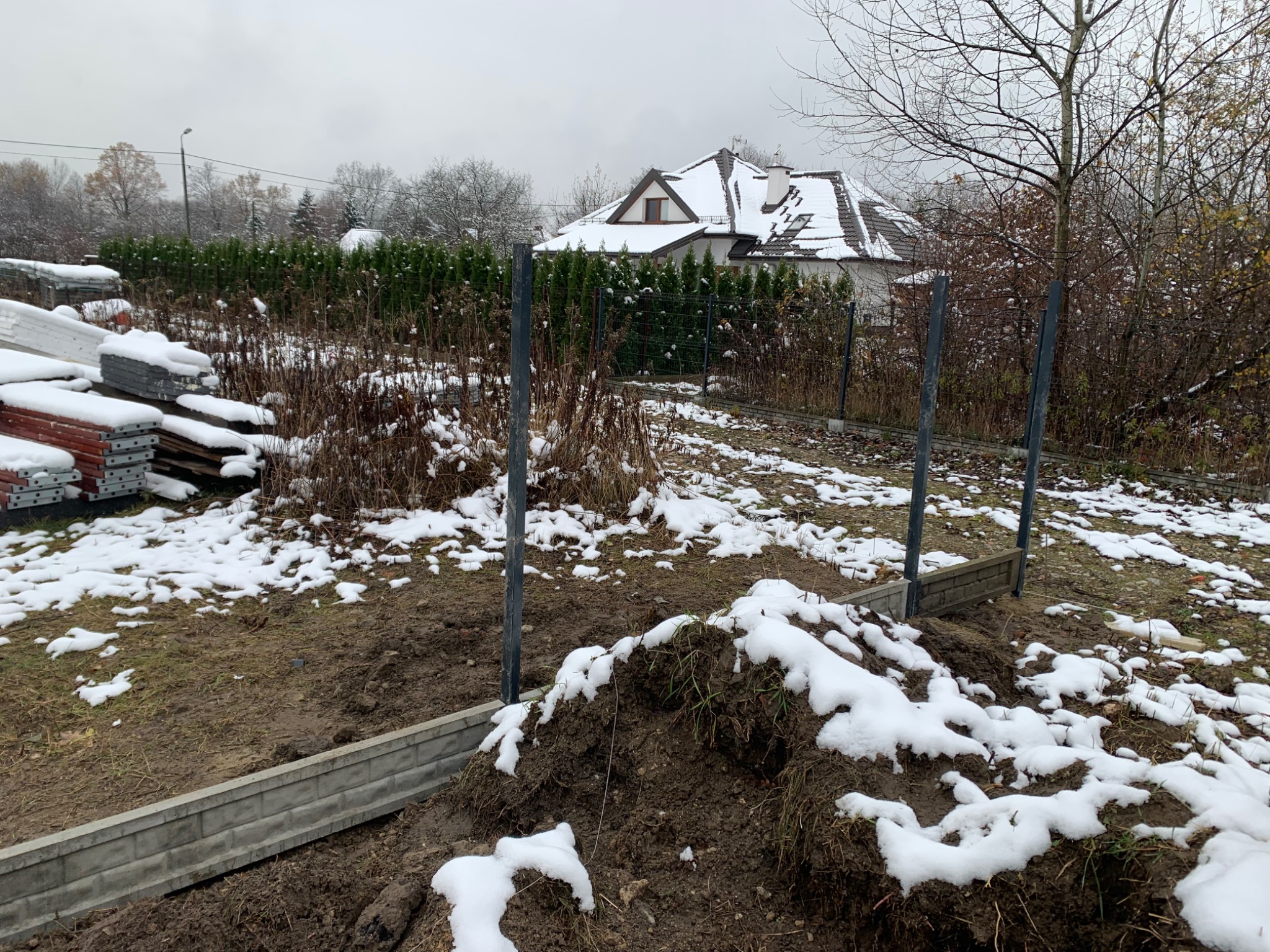 Przygotowanie do montażu ogrodzenia panelowego w zimowej scenerii: widoczne słupki, podmurówka prefabrykowana, ziemia pokryta śniegiem i stosy materiałów budowlanych w tle.