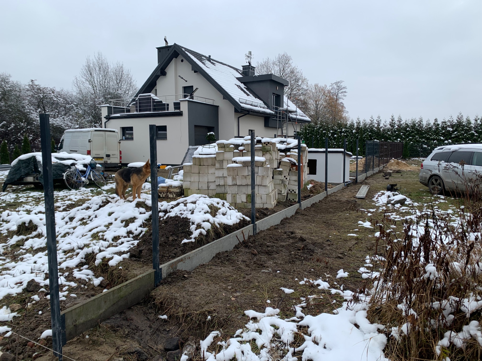 Montaż ogrodzenia panelowego z metalowymi słupkami w zimowej scenerii, na tle ośnieżonego domu i sterty bloczków, z psem owczarkiem niemieckim w kadrze.