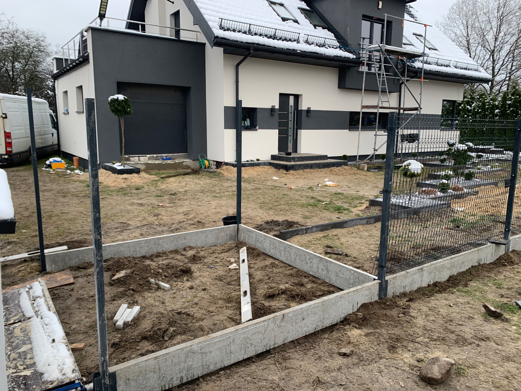 Fundament pod ogrodzenie z siatki na tle nowoczesnego domu z ośnieżonym dachem i rusztowaniem przy elewacji.