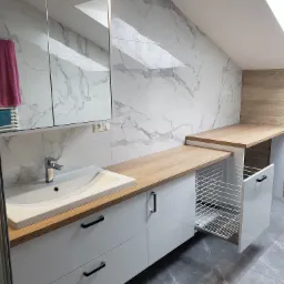 Biała łazienka na poddaszu z marmurową ścianą, umywalką nablatową, drewnianym blatem i wysuwanym koszem na pranie.