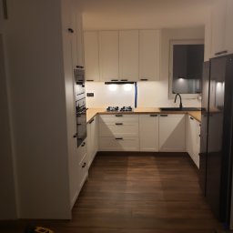 Max Craft - Nowoczesna kuchnia z białymi szafkami, drewnianym blatem i czarnymi uchwytami, widok na kuchenkę gazową i zlew, na podłodze widoczny odkurzacz.