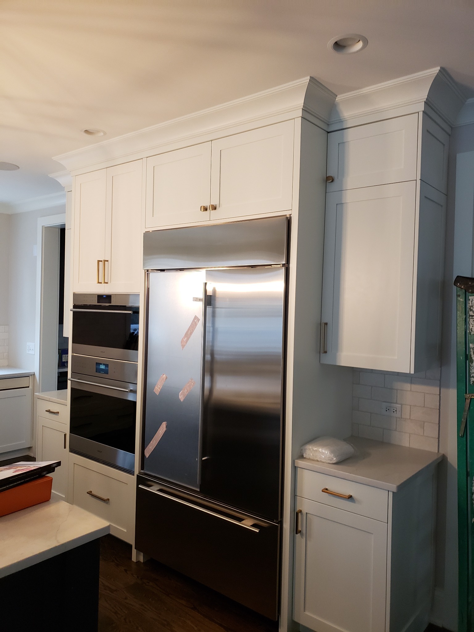 Biała zabudowa kuchenna z górnymi szafkami z dekoracyjnym gzymsem, piekarnik w zabudowie i lodówka ze stali nierdzewnej z folią ochronną na drzwiach.