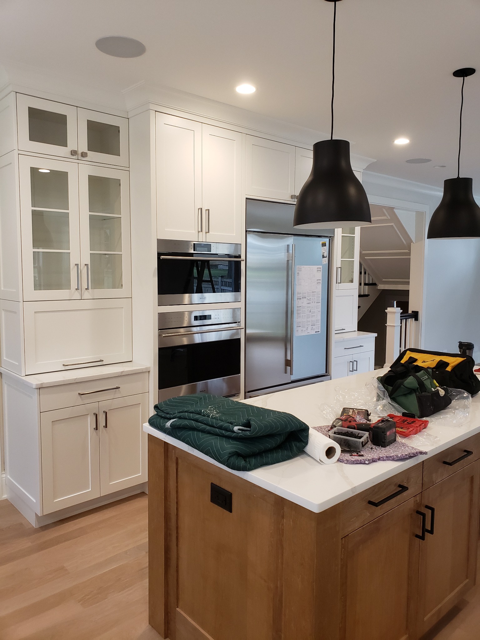 Nowoczesna kuchnia z białymi szafkami, zabudowanymi piekarnikami, lodówką ze stali nierdzewnej i wyspą z drewnianymi frontami, na blacie rozłożone narzędzia i materiały zabezpieczające.