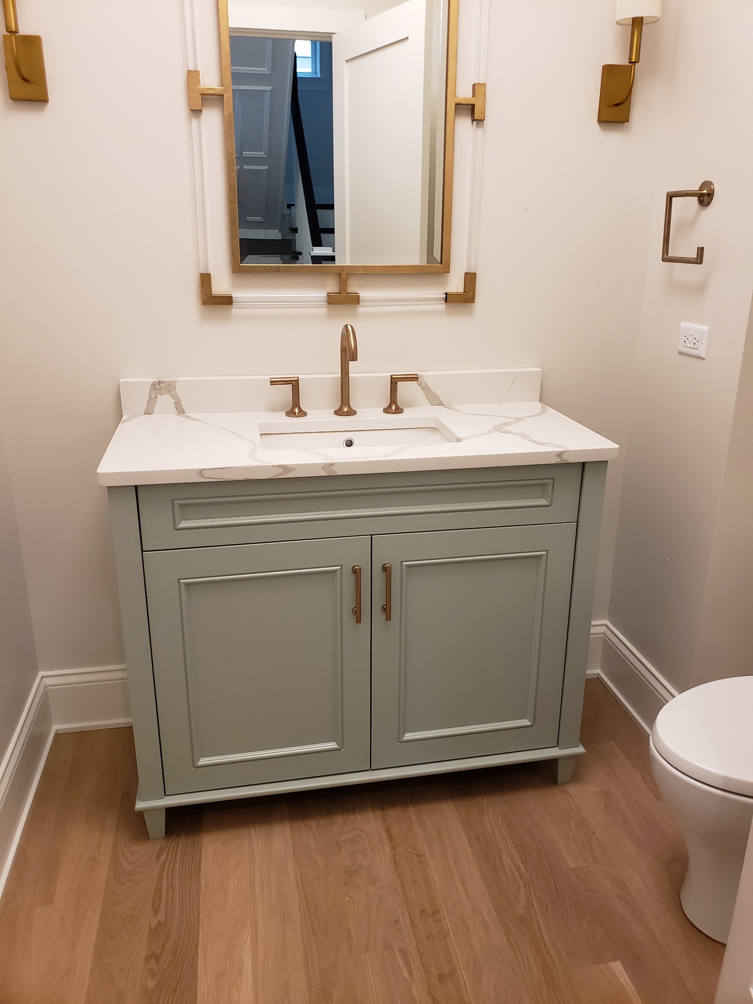 Jasnoszara szafka łazienkowa z blatem z efektem marmuru, złotą armaturą i lustrem z akcentami w kolorze złota, uzupełniona drewnopodobną podłogą i białym sedesem.