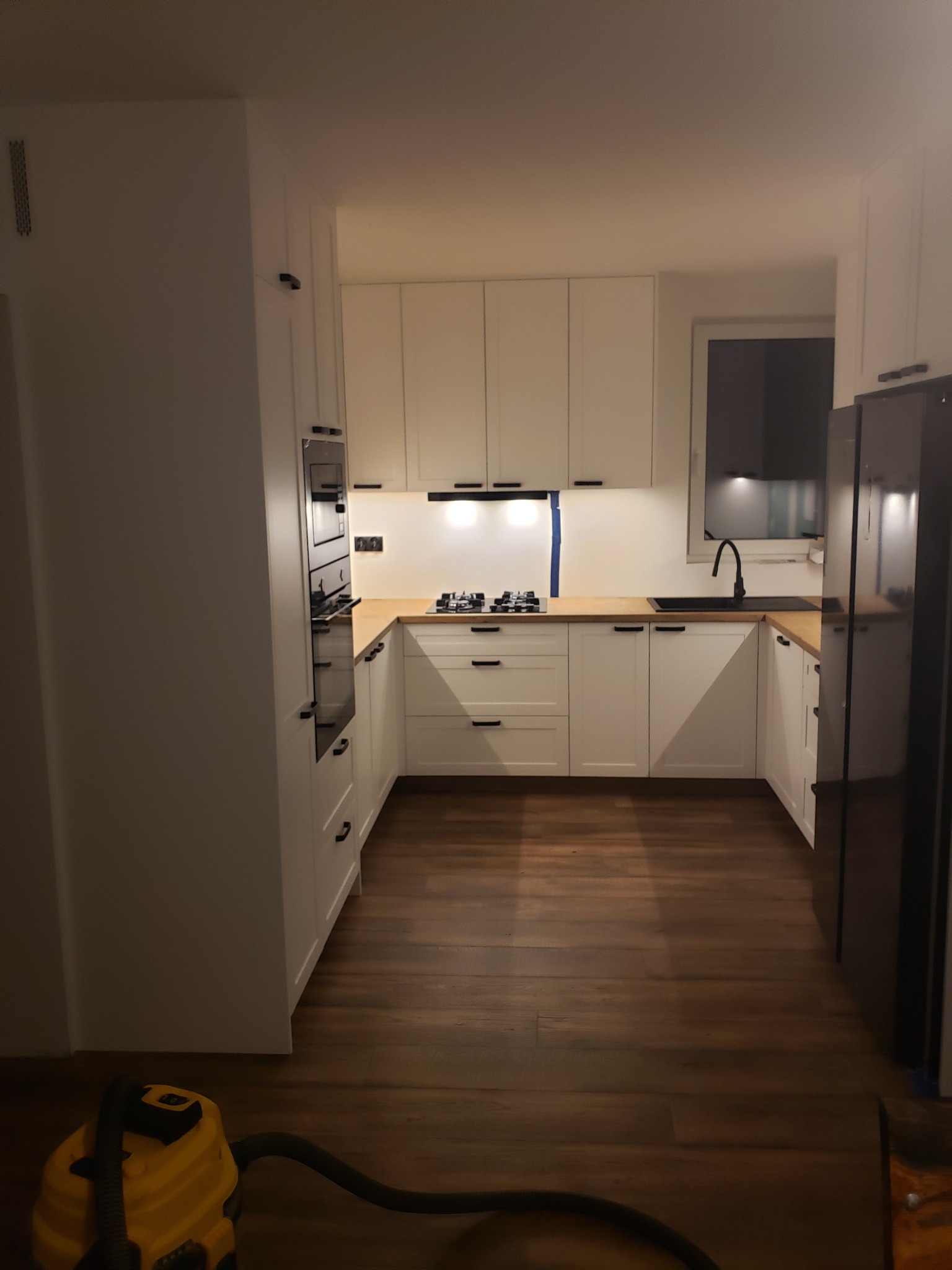 Nowoczesna kuchnia z białymi szafkami, drewnianym blatem i czarnymi uchwytami, widok na kuchenkę gazową i zlew, na podłodze widoczny odkurzacz.