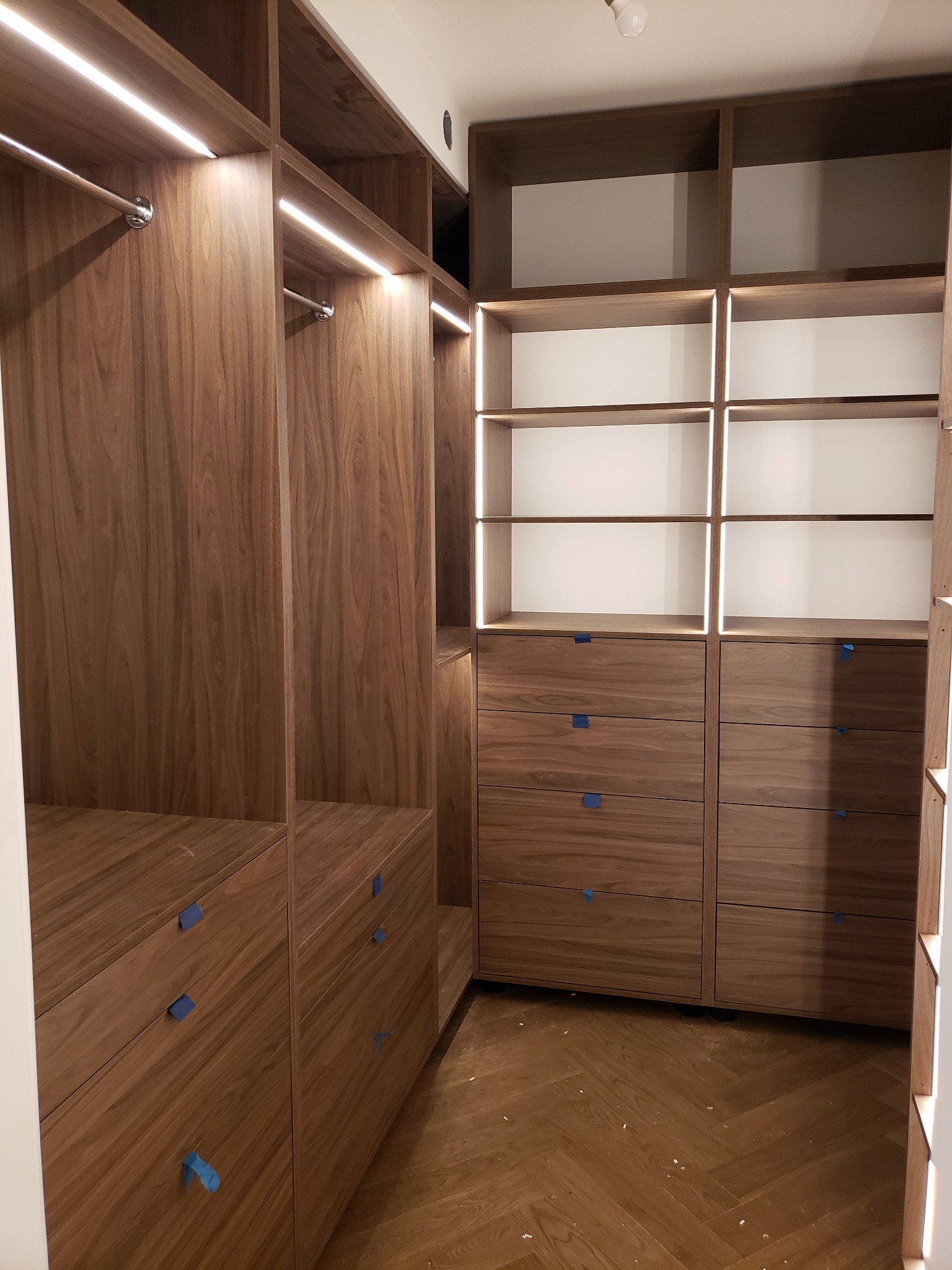 Wnętrze garderoby z drewna orzechowego z oświetleniem LED, widoczne półki, szuflady z niebieską taśmą zabezpieczającą uchwyty, podłoga ułożona w jodełkę.