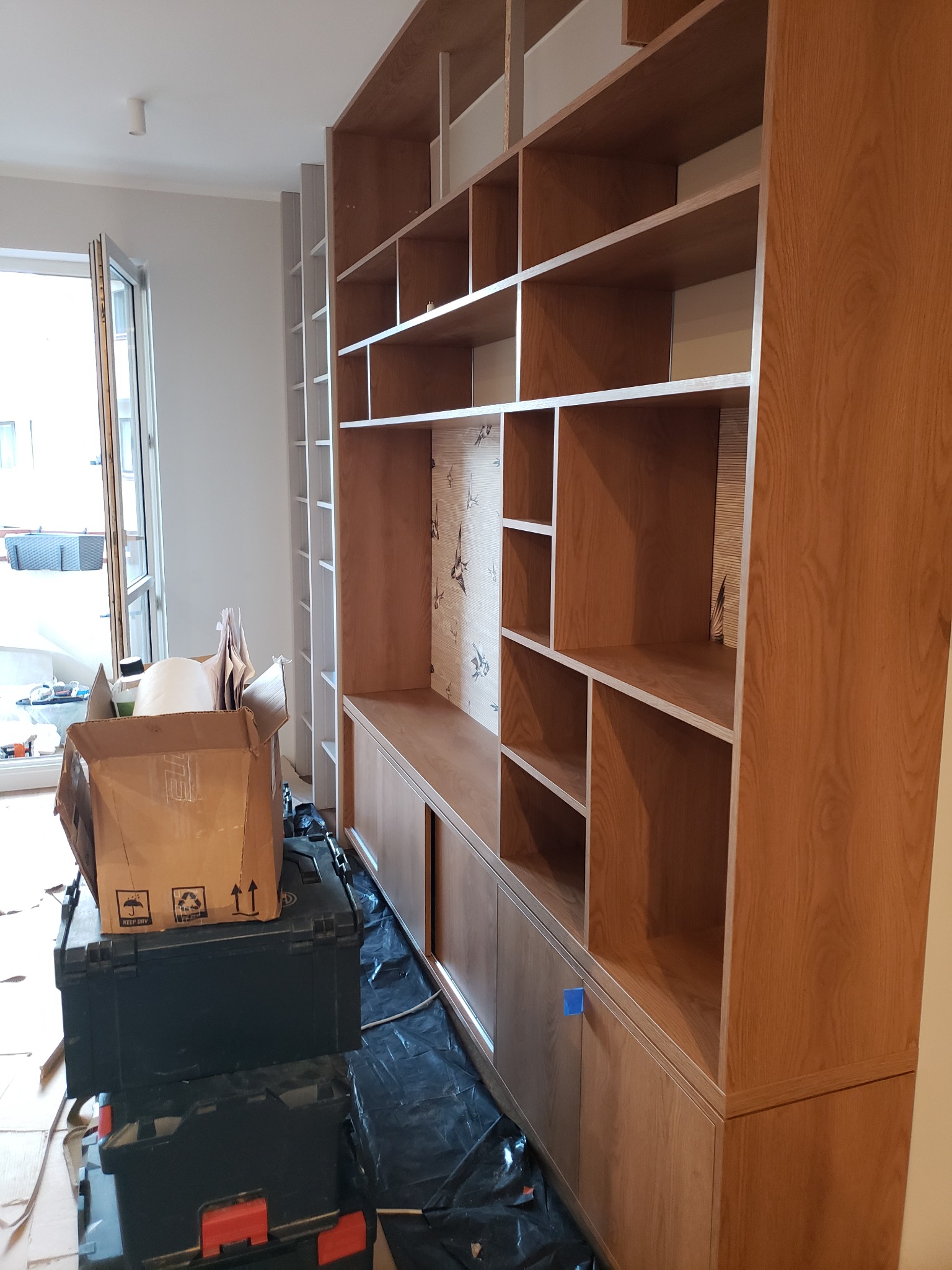 Montaż dużej, drewnianej szafy wnękowej z otwartymi półkami i przesuwnymi drzwiami, widoczne narzędzia i karton, okno w tle, etap prac wykończeniowych.