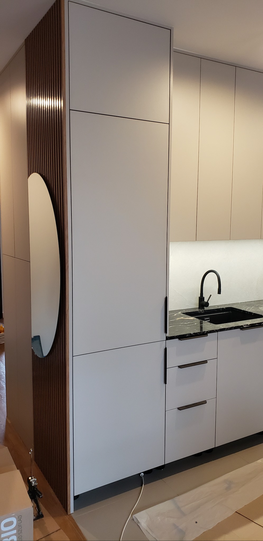 Nowoczesna zabudowa kuchenna z jasnymi frontami, czarnym blatem i okrągłym lustrem wkomponowanym w drewniane lamele. Minimalistyczne uchwyty i oświetlenie LED.