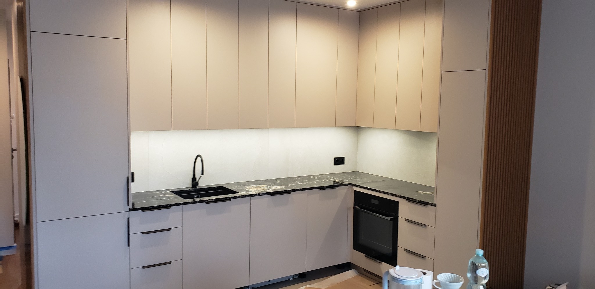 Jasna kuchnia na wymiar z czarnym blatem, minimalistyczny design. Górne szafki podświetlone, widoczny zlew i piekarnik. Nowoczesne wykończenie.